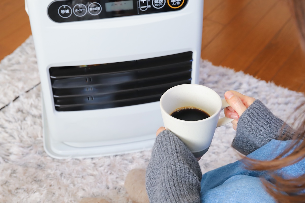 ファンヒーターの前でコーヒーを持ちながら温まる人。