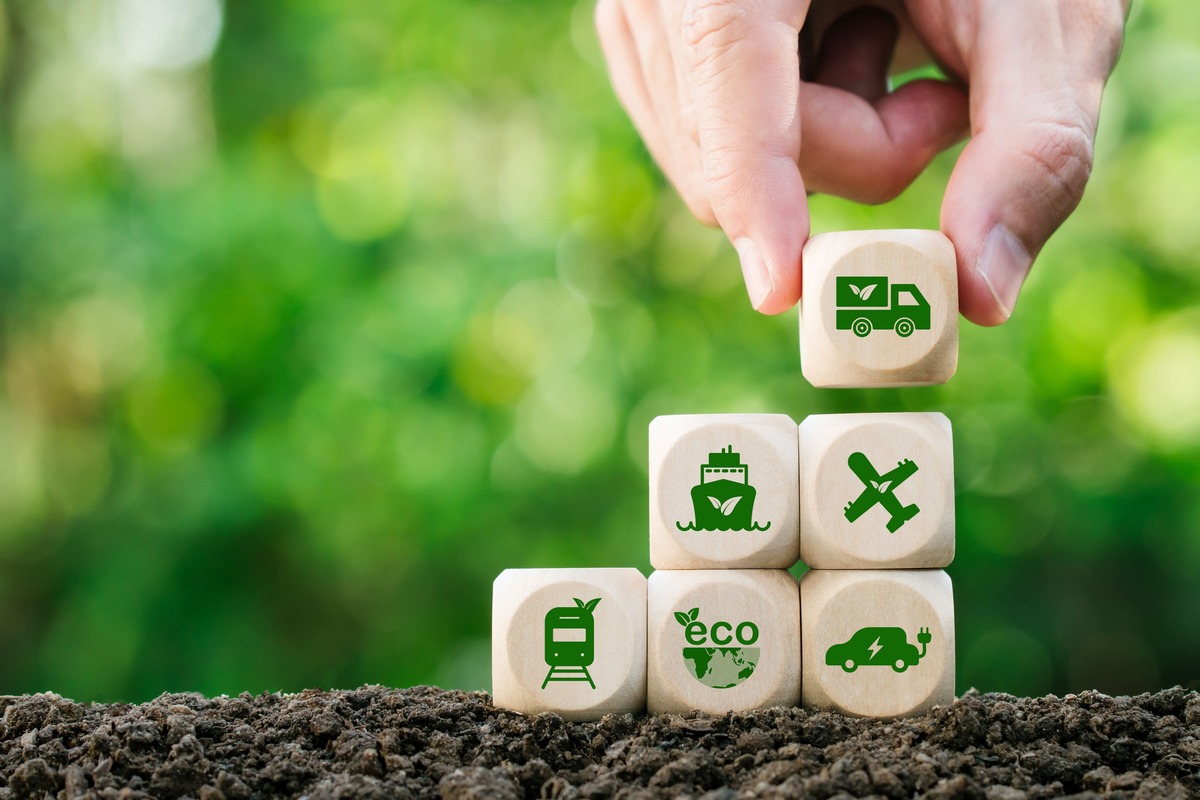 緑背景の土の上に積まれた環境配慮型輸送アイコンの木製ブロック。環境負荷軽減の取り組みを象徴するイメージ画像。