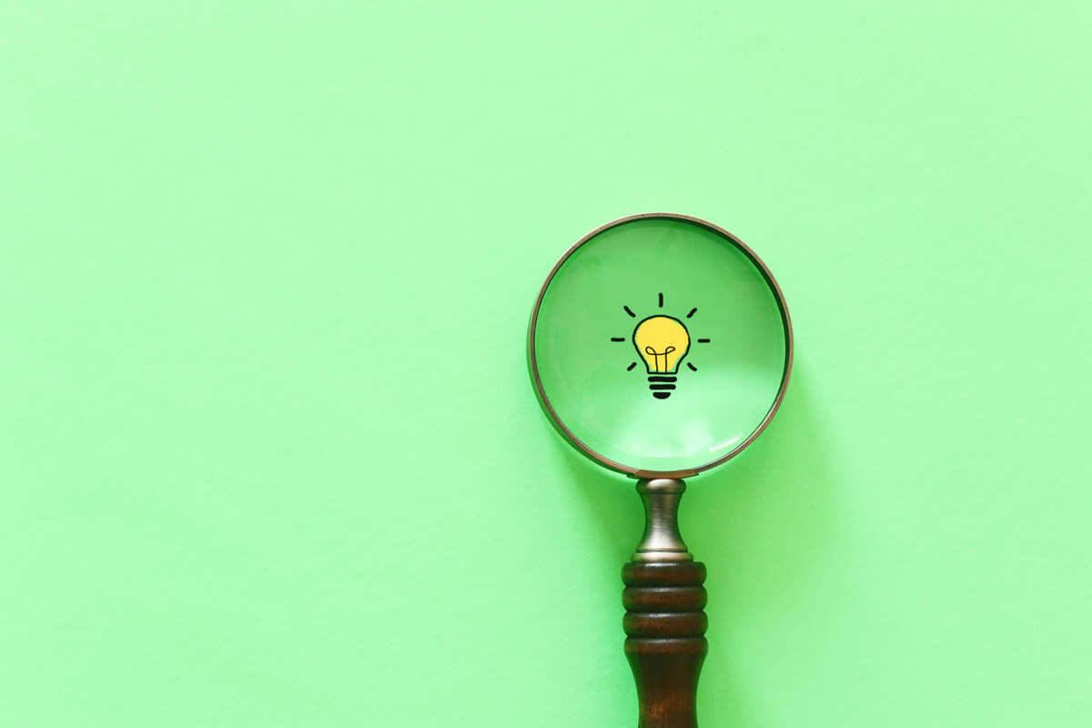 虫眼鏡のレンズに電球のアイコンが映し出されたグリーン背景の画像。電気代の節約には電力会社の切り替えがおすすめであることを示すイメージ画像。