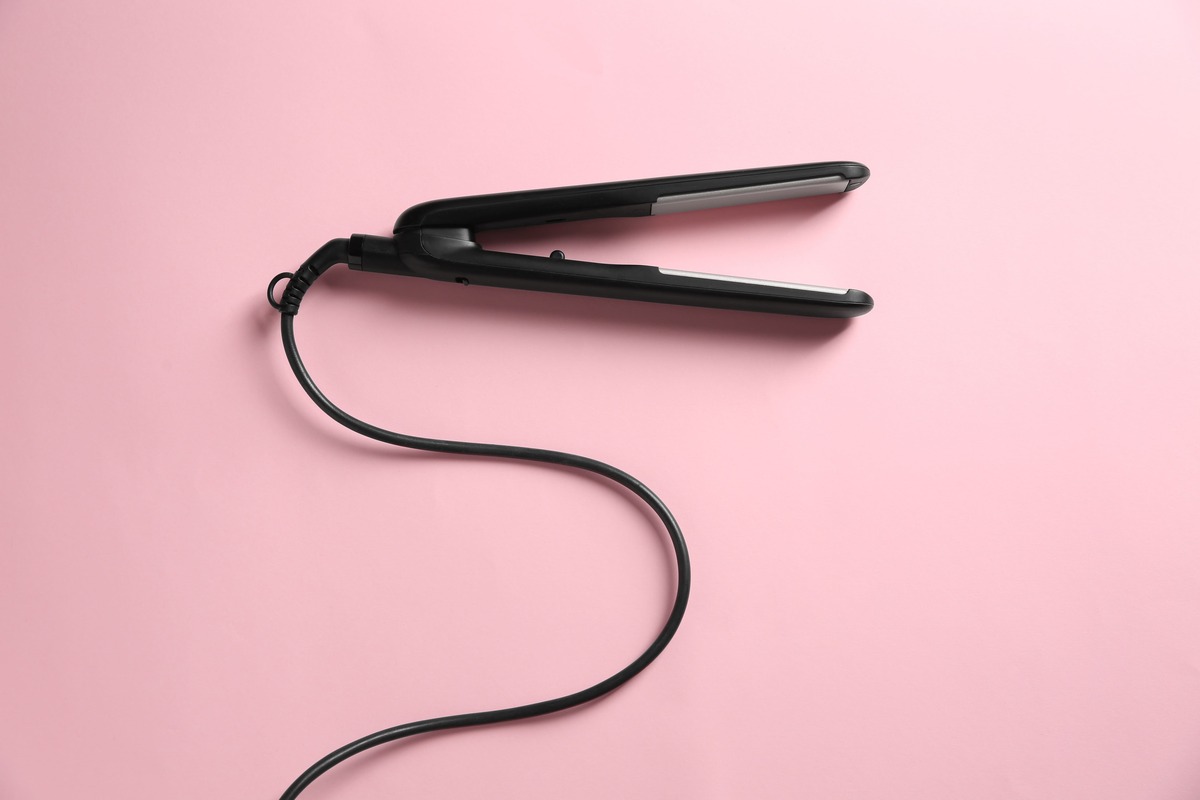 ピンクの背景に置かれた黒いヘアアイロン本体とコード。ヘアアイロンをつけっぱなしにした場合の電気代を表すイメージ。