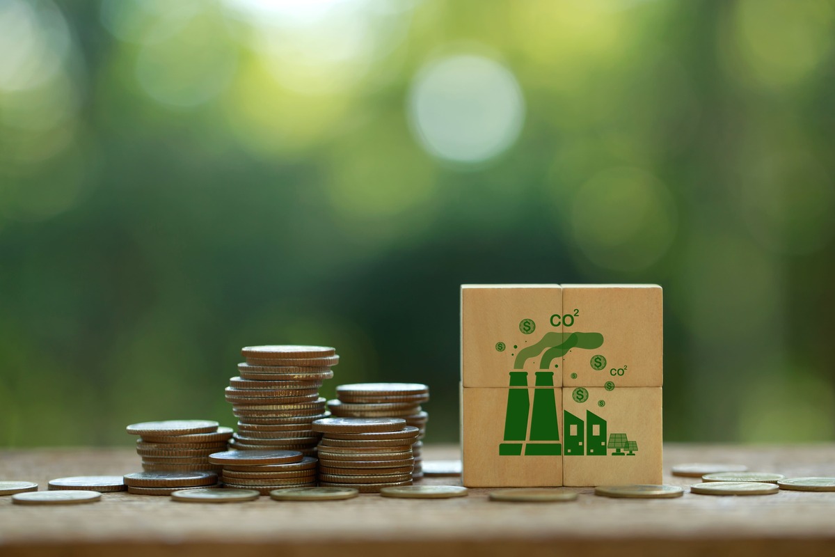 コインの山の隣に置かれた、CO₂・工場・太陽光発電のアイコンが描かれた木製ブロック。コスト面など、カーボンリサイクルの課題を象徴するイメージ。