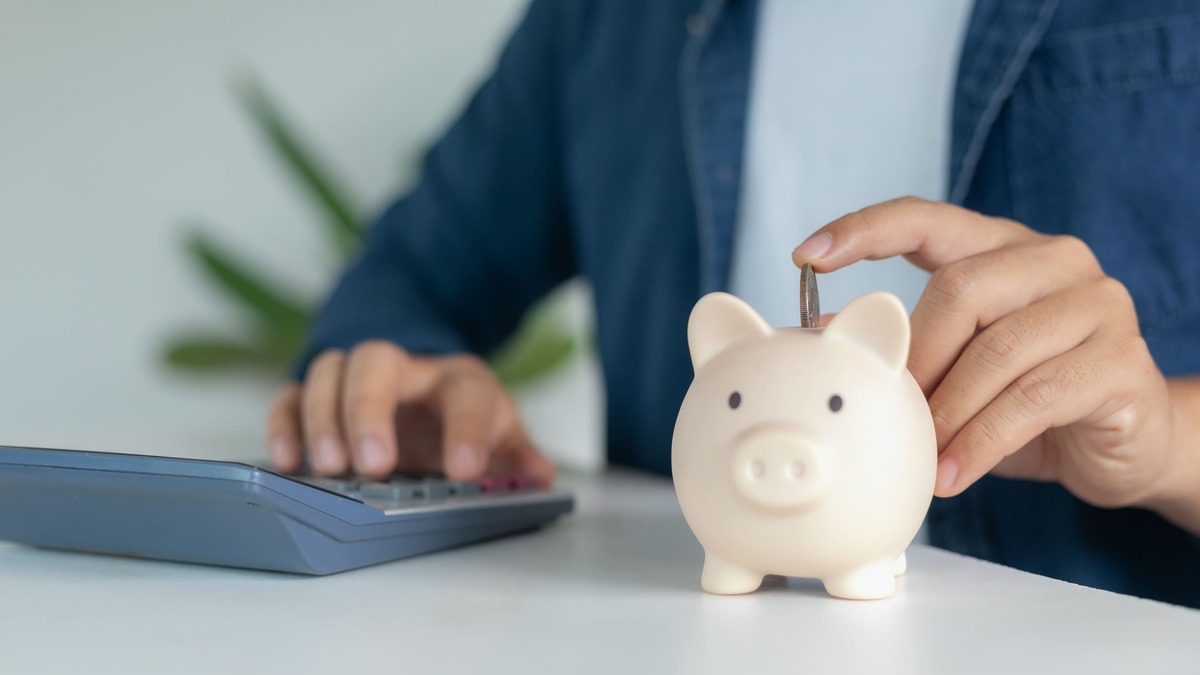 白い豚の貯金箱にコインを入れる手と隣に置かれた電卓。家計の節約を示すイメージ。