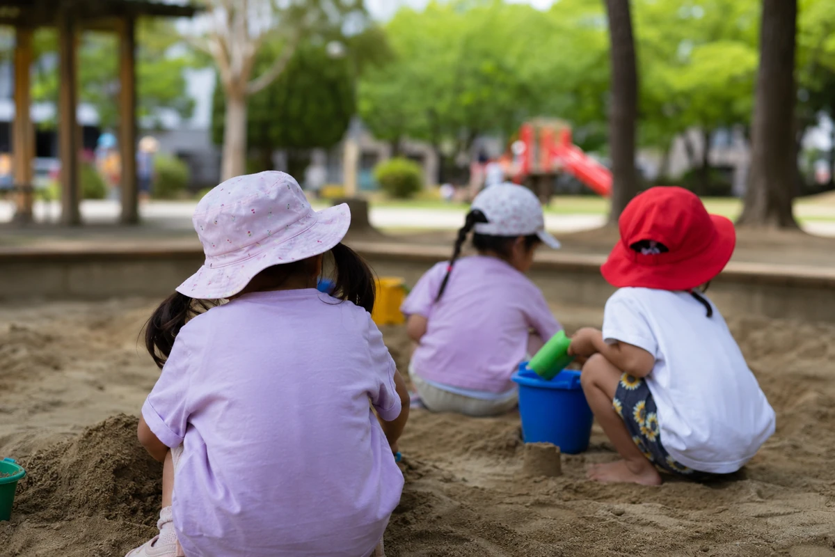 3人の子どもたちが公園の砂場でバケツやスコップを使って遊んでいる後ろ姿。夏の野外遊びと子どもが過ごす環境を示すイメージ写真。