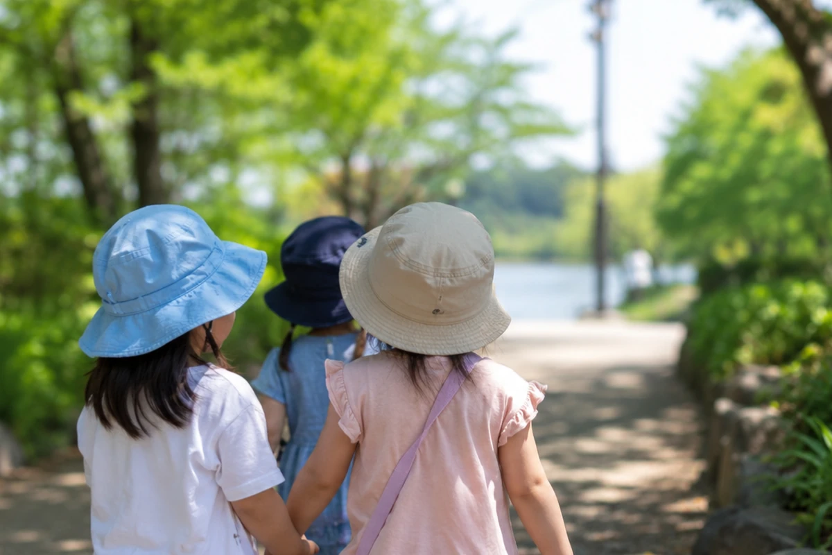 帽子をかぶった3人の子どもが緑豊かな公園の遊歩道の手をつないで歩いている後ろ姿。暑さの和らぐ日陰で過ごすことを示唆するイメージ写真。