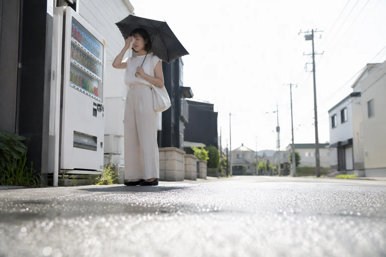 炎天下の路上で日傘をさし、額の汗をぬぐう女性。猛暑による体への負担を表す写真。
