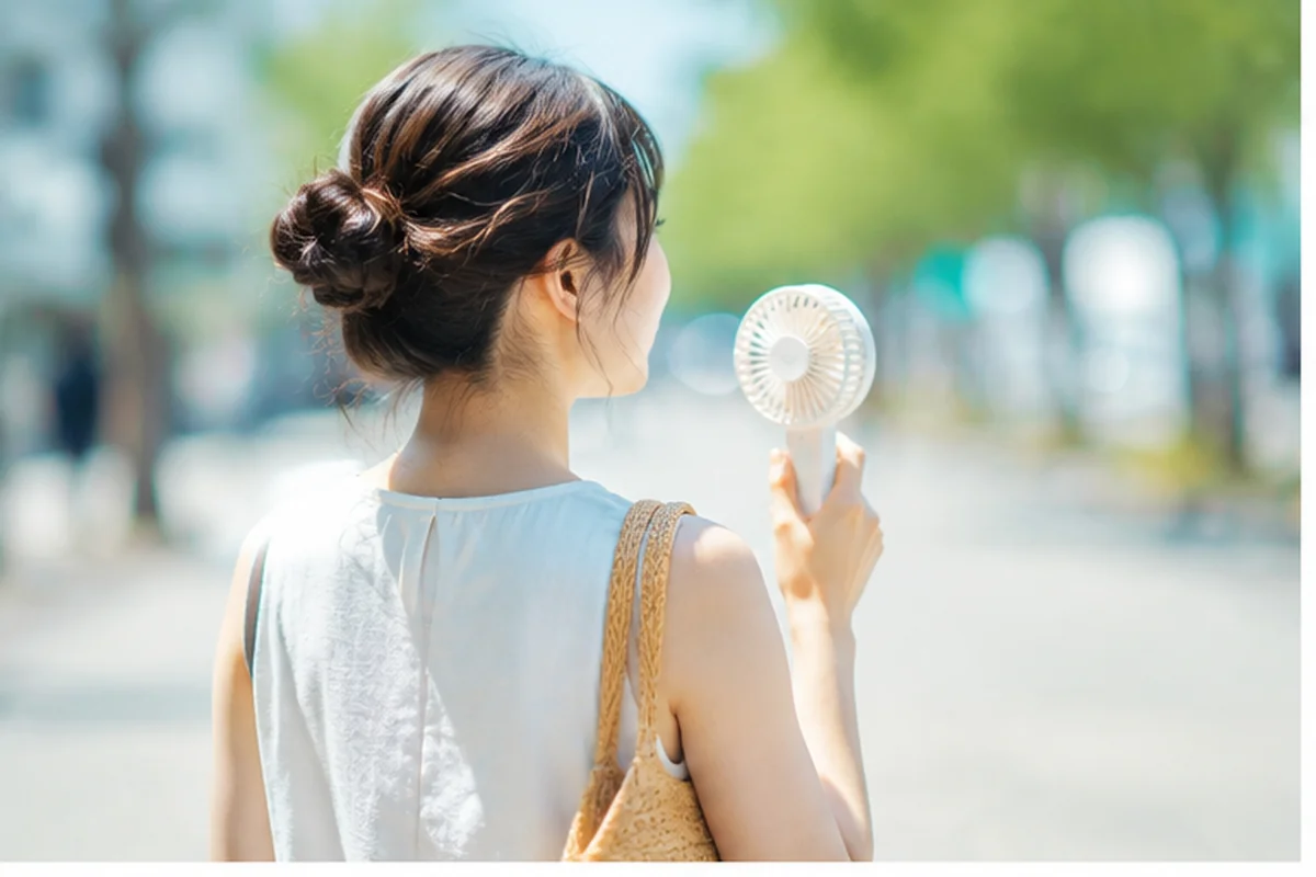 夏の街中でハンディファンを使う女性。夏が長く厳しくなっていることを表す写真。