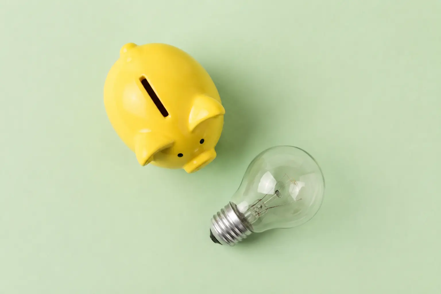 淡い緑の背景に置かれた黄色い貯金箱と電球。電気代の節約を示すイメージ。